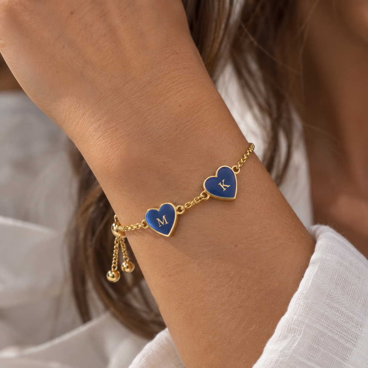 A close-up of a gold chain bracelet worn on a wrist, featuring two heart charms with blue enamel inlays engraved with the letters 'M' and 'K', along with small dangling gold beads.