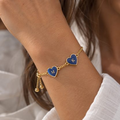 A close-up of a gold chain bracelet worn on a wrist, featuring two heart charms with blue enamel inlays engraved with the letters 'M' and 'K', along with small dangling gold beads.