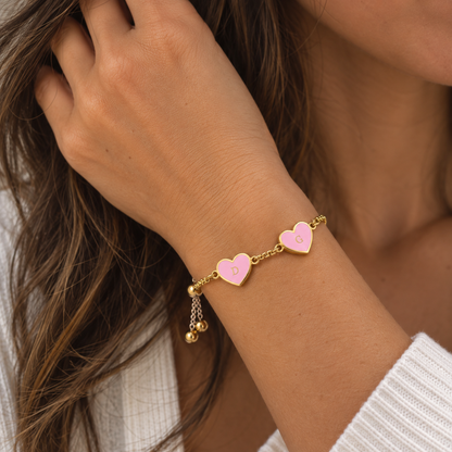 A close-up of a woman's wrist wearing a delicate gold chain bracelet with two pink heart charms, one engraved with the letter 'D' and the other with 'G', along with small dangling gold beads.