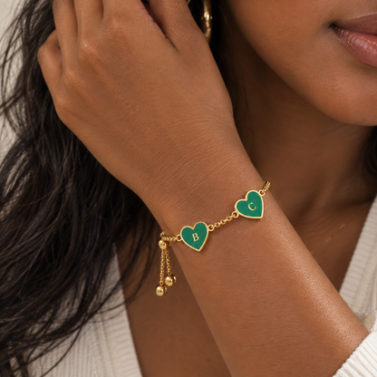 A close-up of a woman's wrist wearing a gold chain bracelet with two green heart charms, one engraved with the letter 'B' and the other with 'C'.