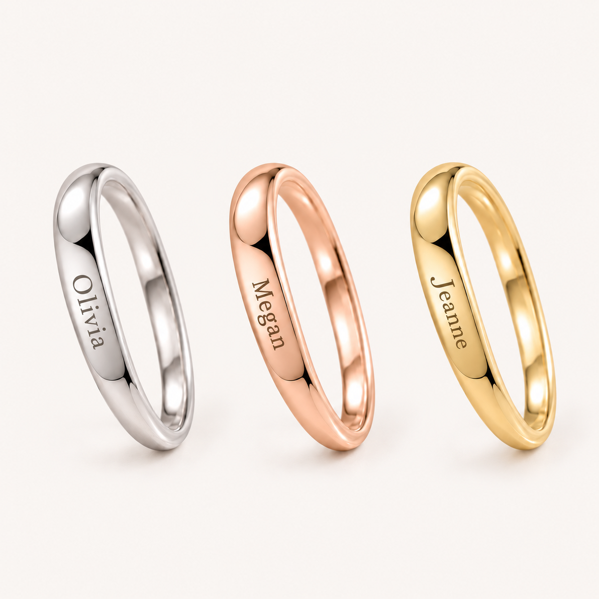 Three slim polished rings on a light warm background: a silver ring engraved 'Olivia', a rose gold ring engraved 'Megan', and a yellow gold ring engraved 'Jeanne'.