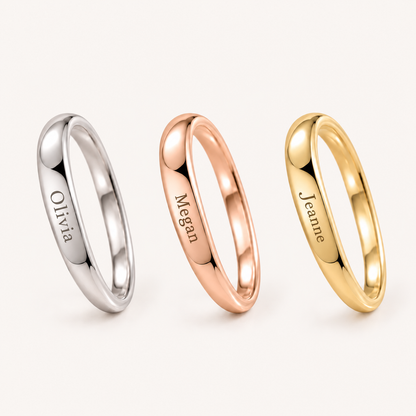 Three slim polished rings on a light warm background: a silver ring engraved 'Olivia', a rose gold ring engraved 'Megan', and a yellow gold ring engraved 'Jeanne'.