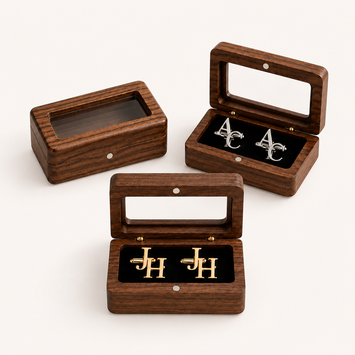 Three rectangular wooden jewelry boxes with glass lids are arranged on a light neutral background. Two boxes are open, displaying black velvet inserts with silver and gold monogram earrings, and one box is closed showing its interior.
