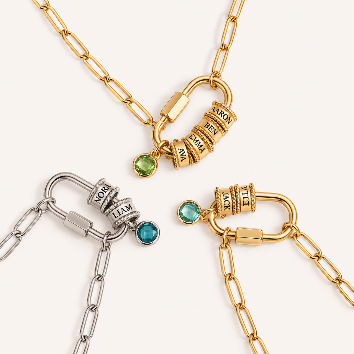 Three personalized locket necklaces with paperclip chains are arranged on a light cream background. Two are gold-toned and one is silver-toned, featuring a combination lock clasp with engraved names (such as 'MOR', 'TEN', 'BEN', 'IVY') and small round gemstone charms in green and blue.