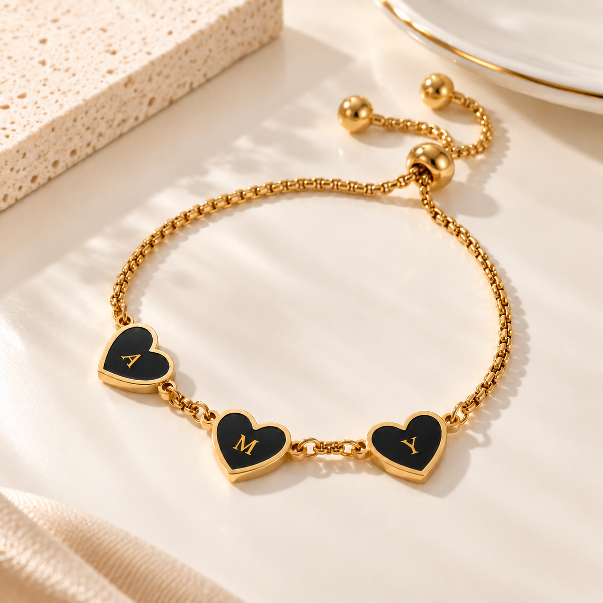 A gold chain bracelet with three heart charms, each featuring black enamel and a gold monogram letter (A, M, Y), displayed on a light beige surface with soft shadows.