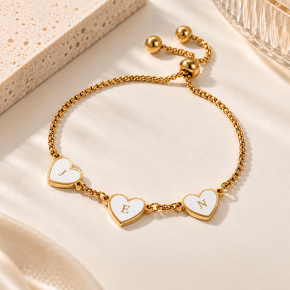 A gold-tone adjustable chain bracelet with three white heart charms outlined in gold, each engraved with a letter (J, E, N), displayed on a warm cream surface with soft shadows.