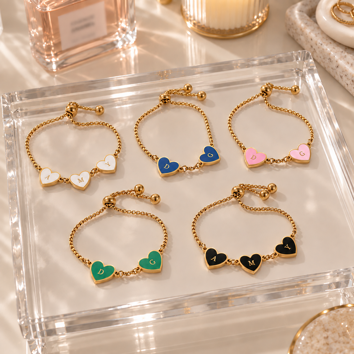 Six adjustable gold chain bracelets with heart charms are arranged on a clear acrylic tray. The heart charms are in different colors—white, blue, pink, green, black, and gold-toned—and each has a small letter engraved on it.