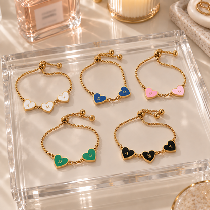 Six adjustable gold chain bracelets with heart charms are arranged on a clear acrylic tray. The heart charms are in different colors—white, blue, pink, green, black, and gold-toned—and each has a small letter engraved on it.