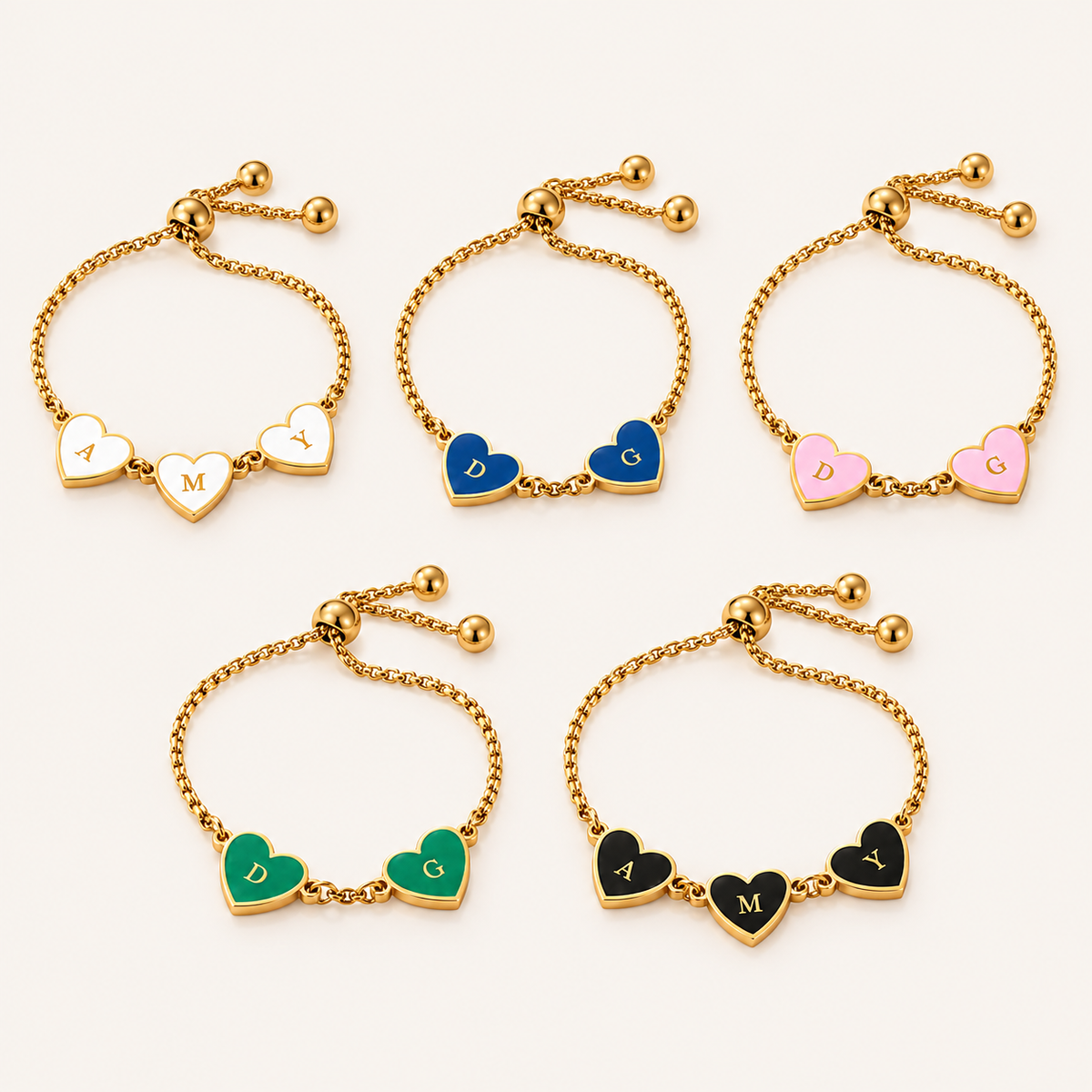 Five adjustable gold-tone chain bracelets with heart charms, arranged on a light beige background. The bracelets feature heart charms in different colors: two white with gold lettering, one blue, one pink, one green, and one black with gold lettering.