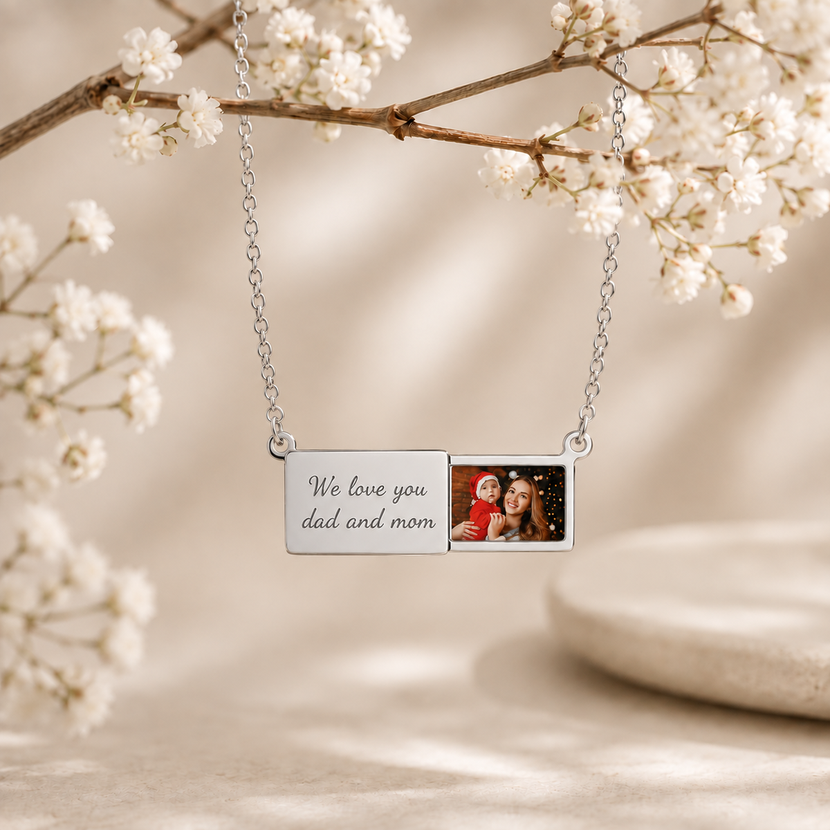 A silver rectangular photo necklace pendant hanging from a delicate chain, with one side engraved with the words 'We love you dad and mom' and the other side showing a small printed photo of two people. The pendant is styled against a soft beige background with white blossoms and a stone.