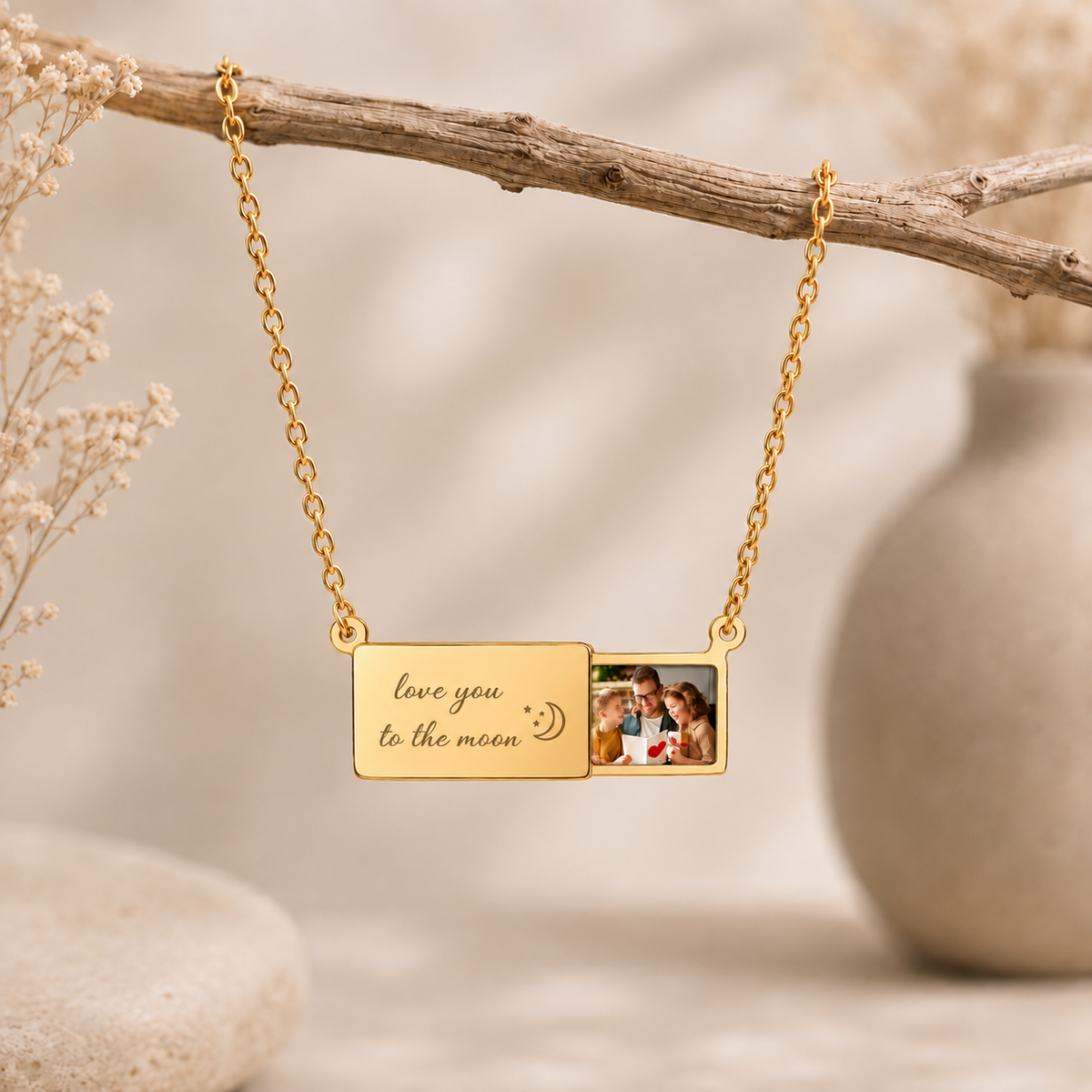 A gold-toned necklace with a rectangular photo pendant hangs from a branch in a softly styled setting. One side of the pendant is engraved with 'love you to the moon' and a crescent moon, while the other side shows a small inset photo of a couple.