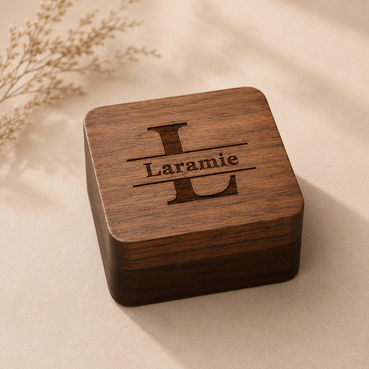 A square wooden keepsake box with rounded corners, featuring an engraved monogram 'L' and the name 'Laramie' on the lid, set on a beige surface with soft shadows and dried floral accents.