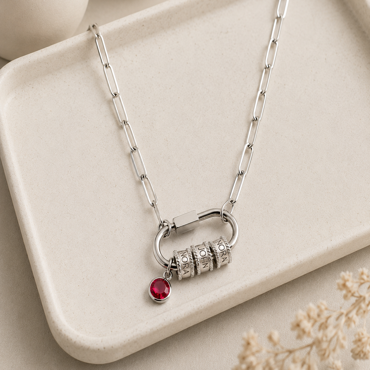 A silver-toned chain necklace with a ring-shaped pendant, three cylindrical charm beads, and a small dangling red gemstone, displayed on a light beige tray.